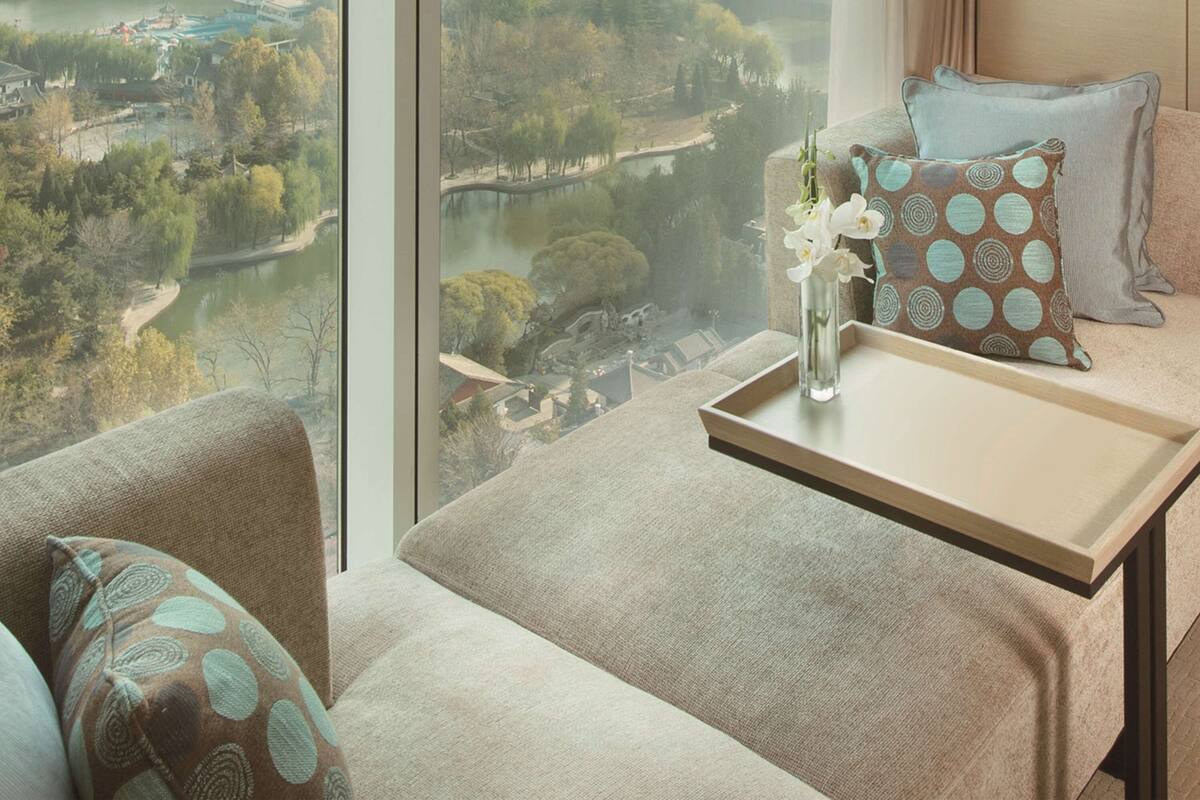 Ett modernt hotellrum med ett stort fönster som erbjuder utsikt över staden, ett litet bord med en vas med blommor och en soffa med mönstrade kuddar.