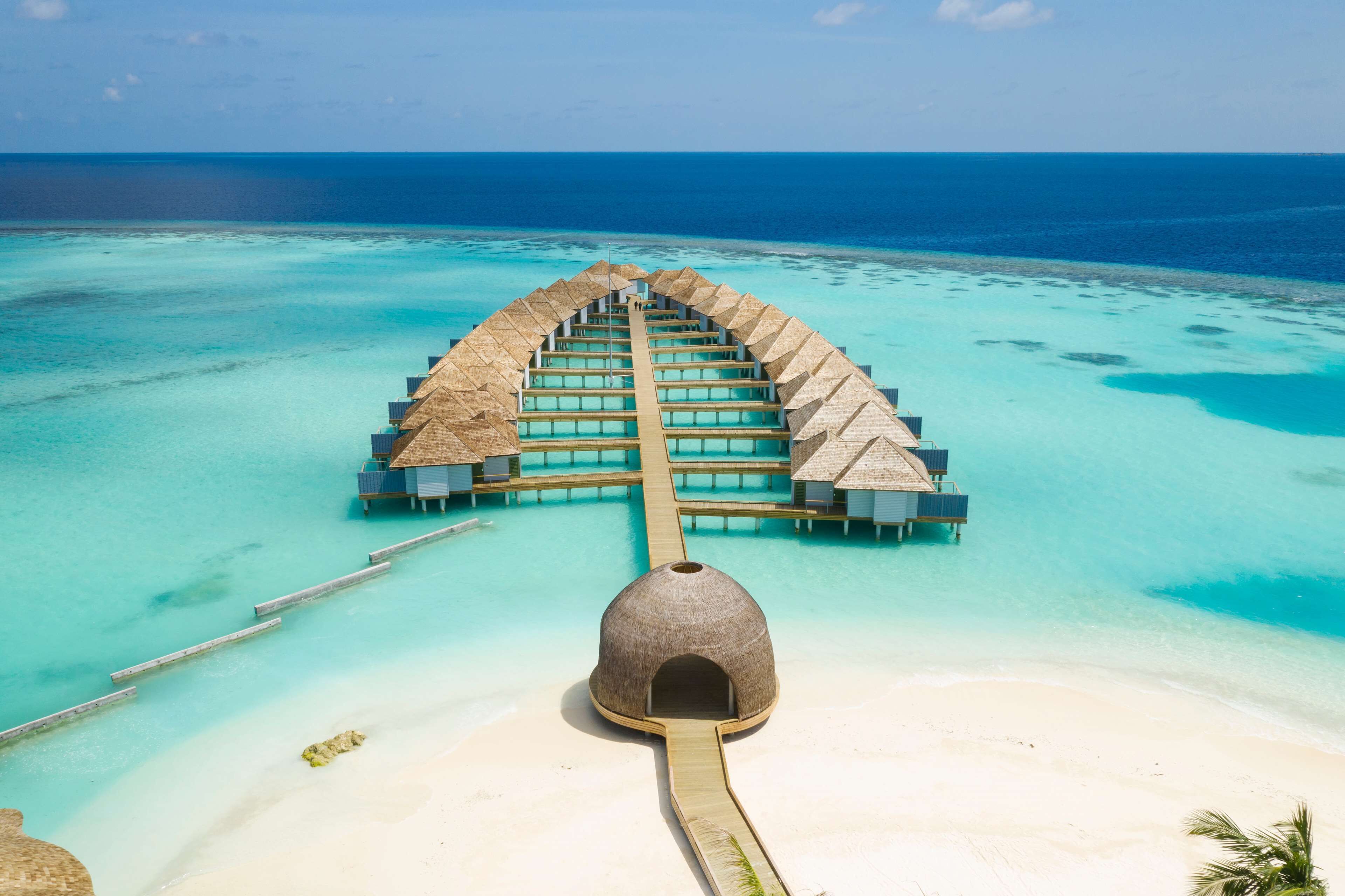 An overwater resort with a central building, connected by walkways, and a beach in the foreground.