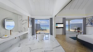 A modern hotel bathroom with a large bathtub, a sink, and a mirror. There is a view of a pool and a balcony with a table and chairs.