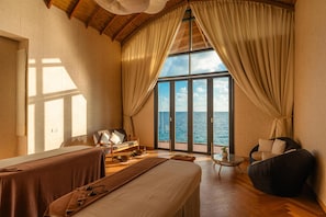 A room with a bed, a sofa, a small table, and a chair, with a view of the sea through large windows. - OUTRIGGER Maldives Maafushivaru Resort (Maafushivaru)