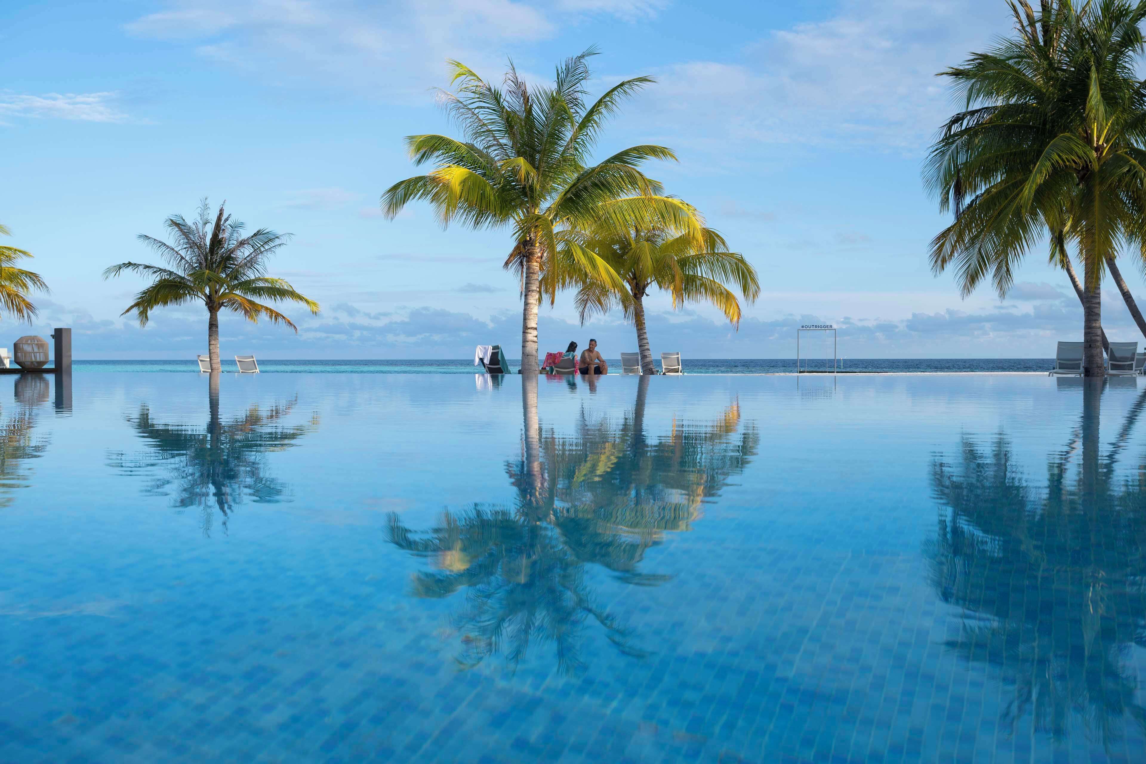 An infinity pool with palm trees and lounge chairs, a clear sky, and the ocean in the background.