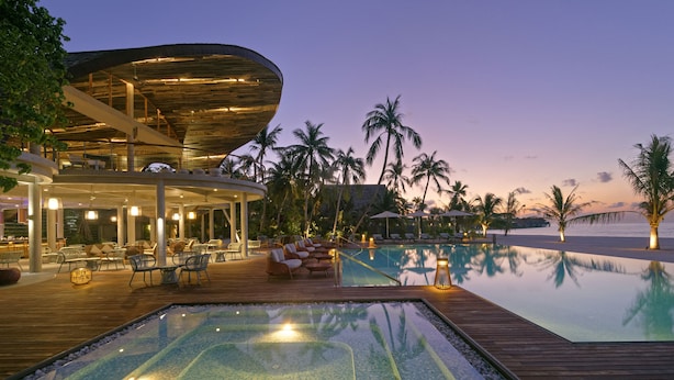 Un resort con una amplia piscina infinita, terraza de madera, zona de descanso y palmeras.