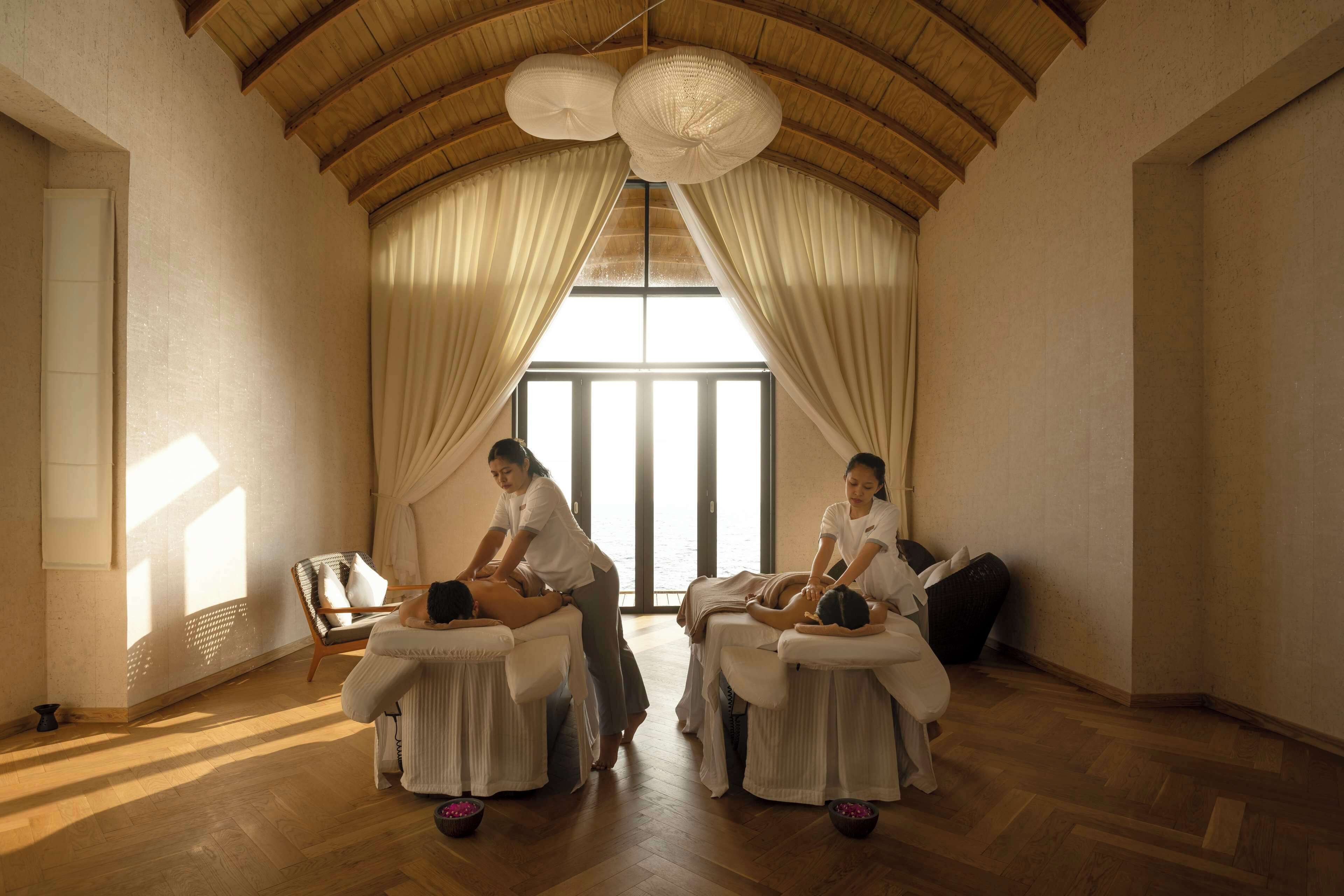 A room with wooden flooring, a high ceiling, and large windows with curtains. There are massage tables with clients and a massage therapist.