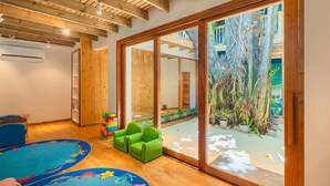 A room with wooden beams, a large sliding glass door to a garden, a blue play mat, and green chairs.