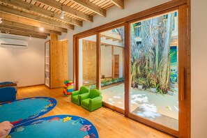 A room with wooden beams, a large sliding glass door to a garden, a blue play mat, and green chairs.