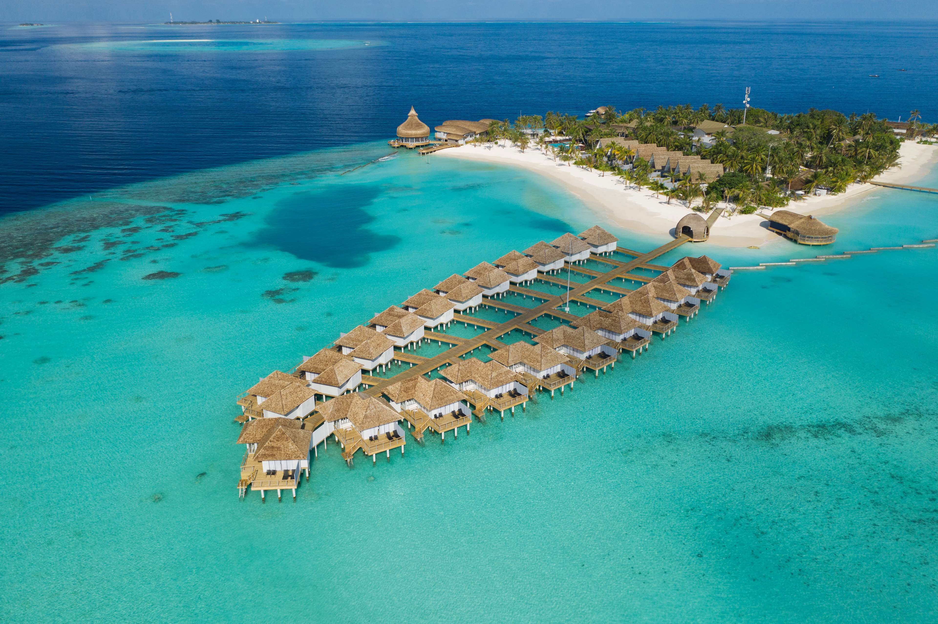 An overwater resort with thatched-roof bungalows connected by walkways, a beach, and palm trees.