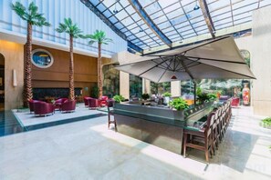 A spacious indoor area with a glass ceiling, a central bar with seating, and palm trees. - Hotel de Royce (Shaoguan)
