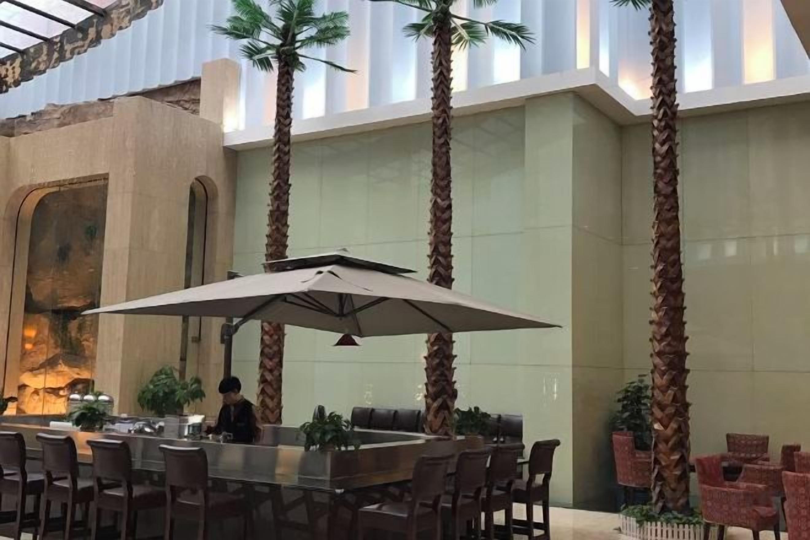 An indoor dining area with a high ceiling, palm trees, and a large umbrella table.