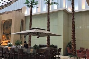 An indoor dining area with a high ceiling, palm trees, and a large umbrella table. - Hotel de Royce (Shaoguan)