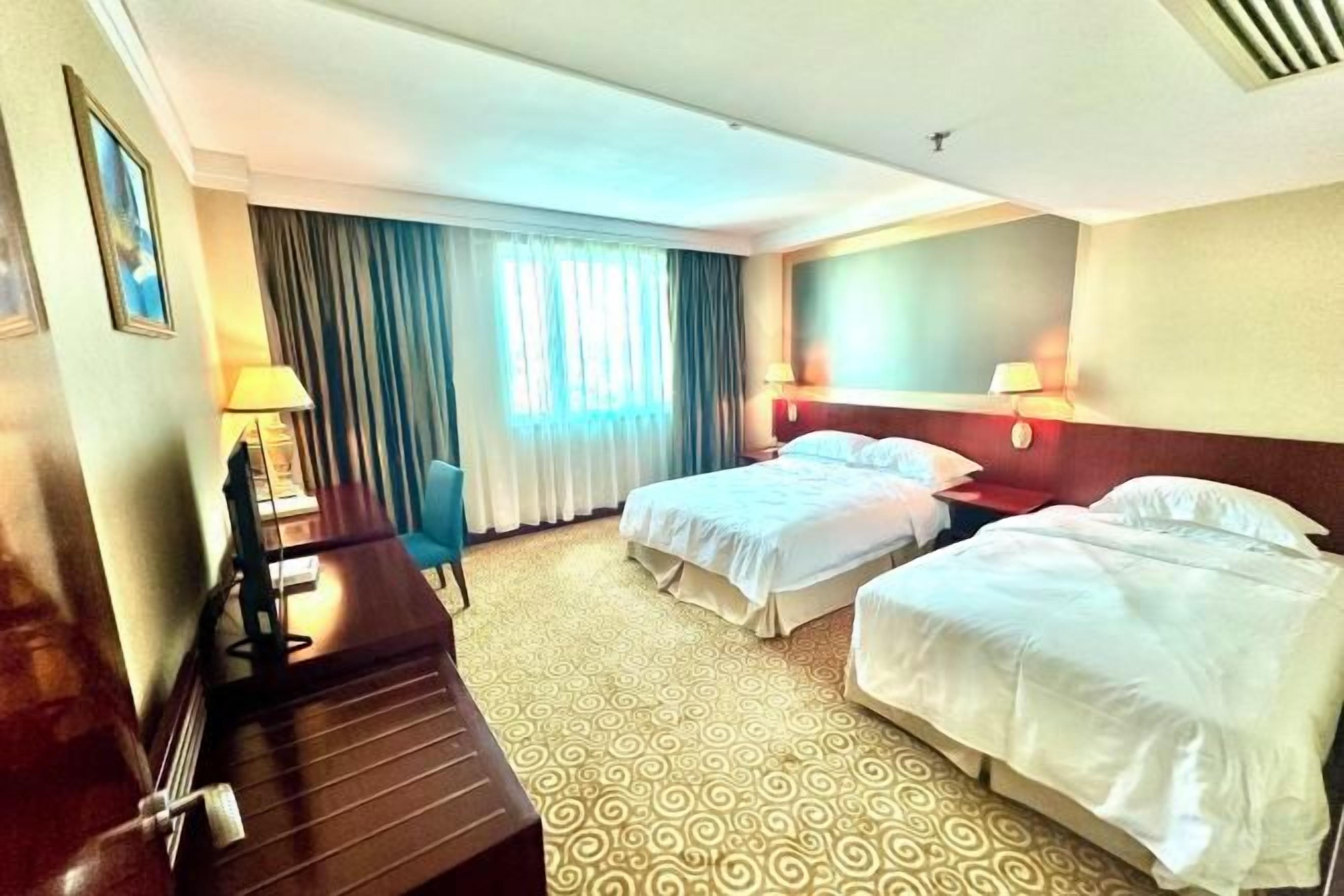 A hotel room with two beds, a desk, a chair, a desk lamp, a nightstand, and a window with curtains.