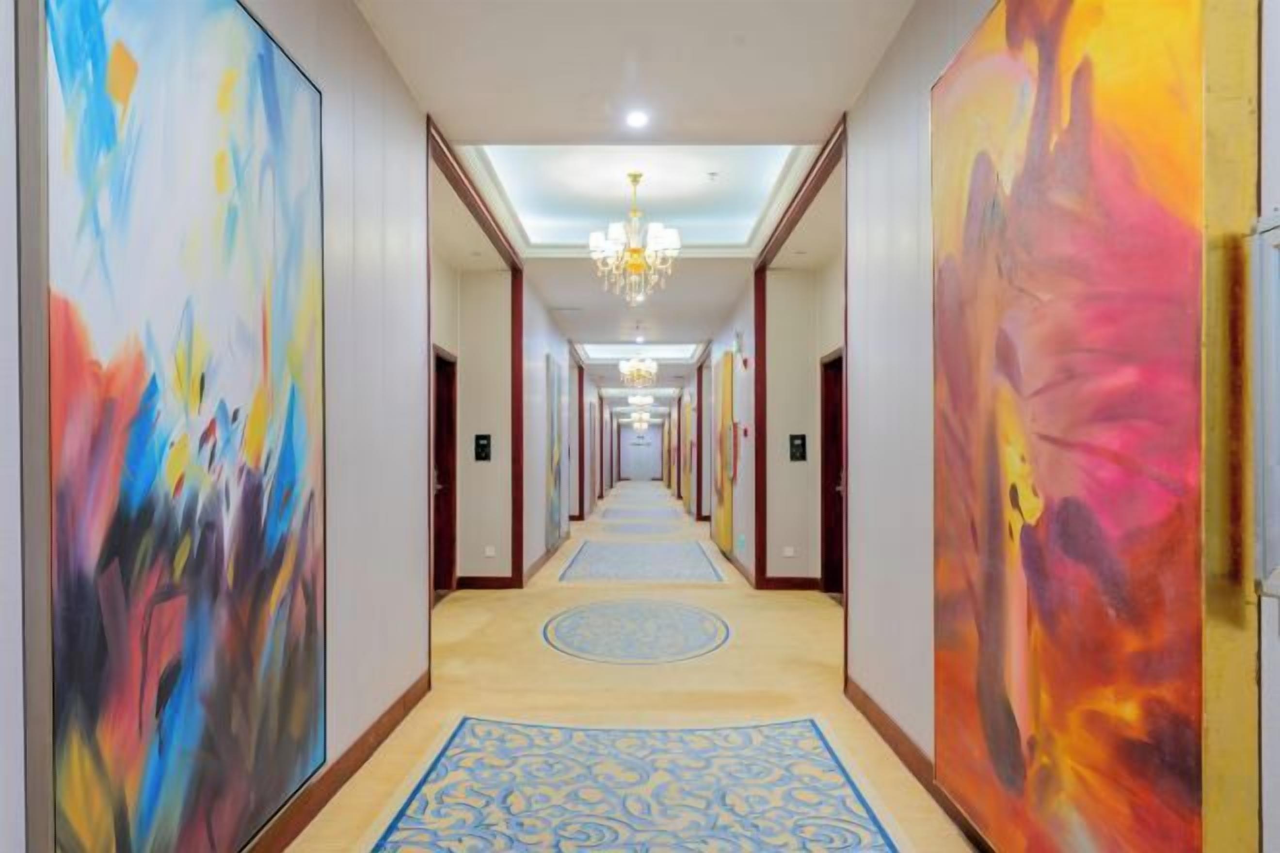 A hotel corridor with a chandelier, blue carpet, and large abstract paintings on the walls.