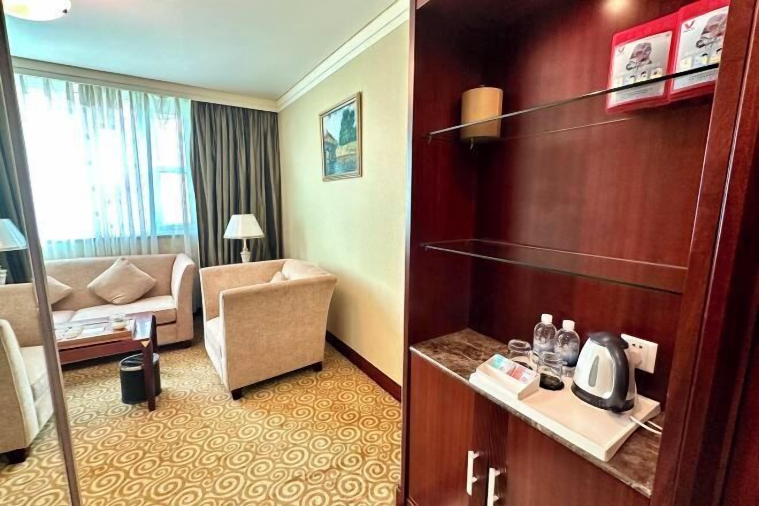 A hotel room with a sofa, armchair, patterned carpet, wooden wardrobe, and a tray with a kettle.