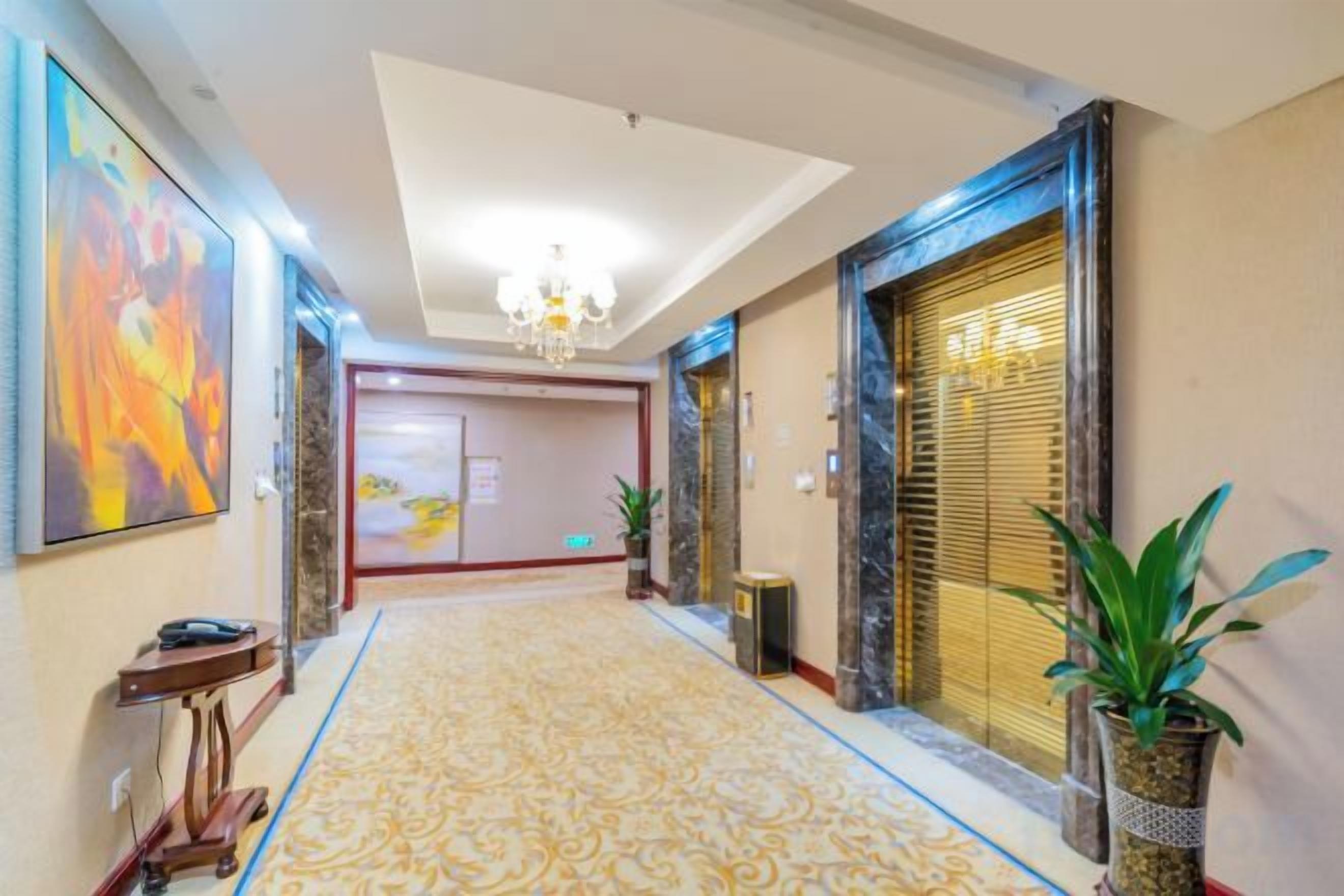 A hotel hallway with a patterned carpet, a chandelier, and a large painting on the wall.