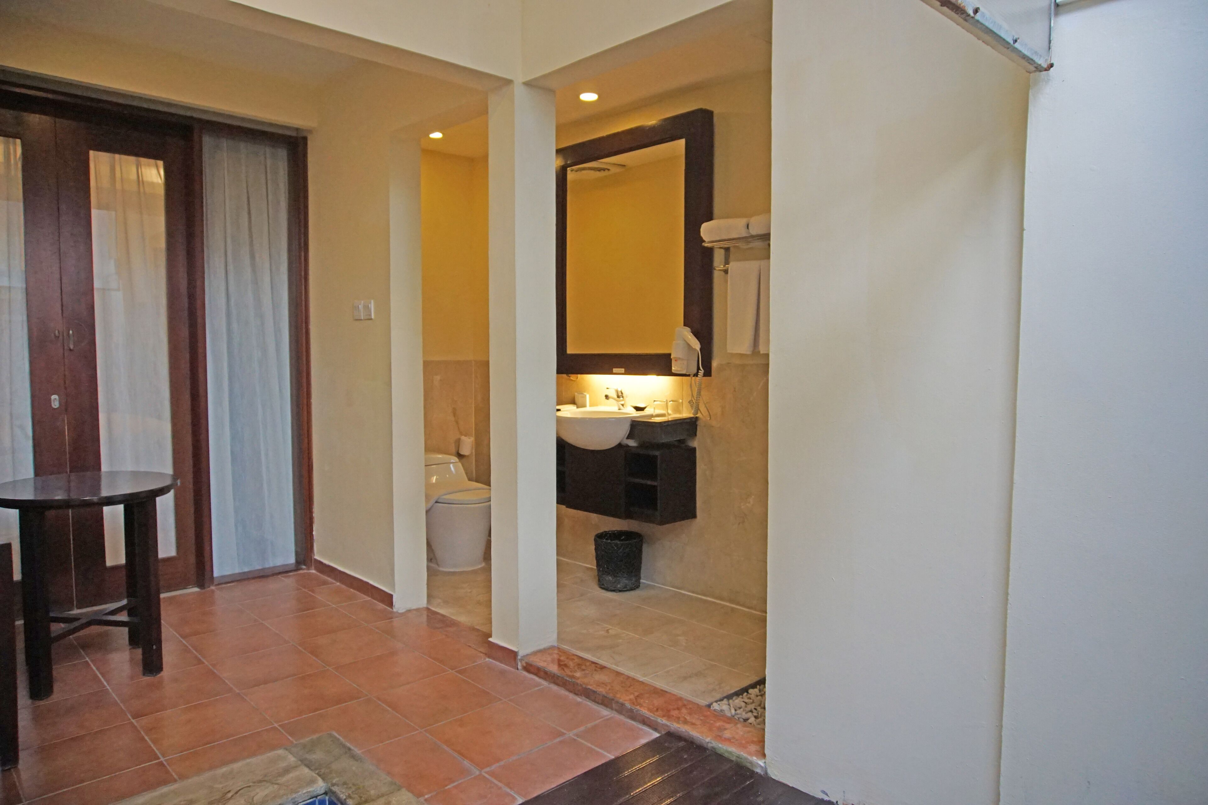 A bathroom with a toilet, sink, and mirror. There is a small table and a trash can.