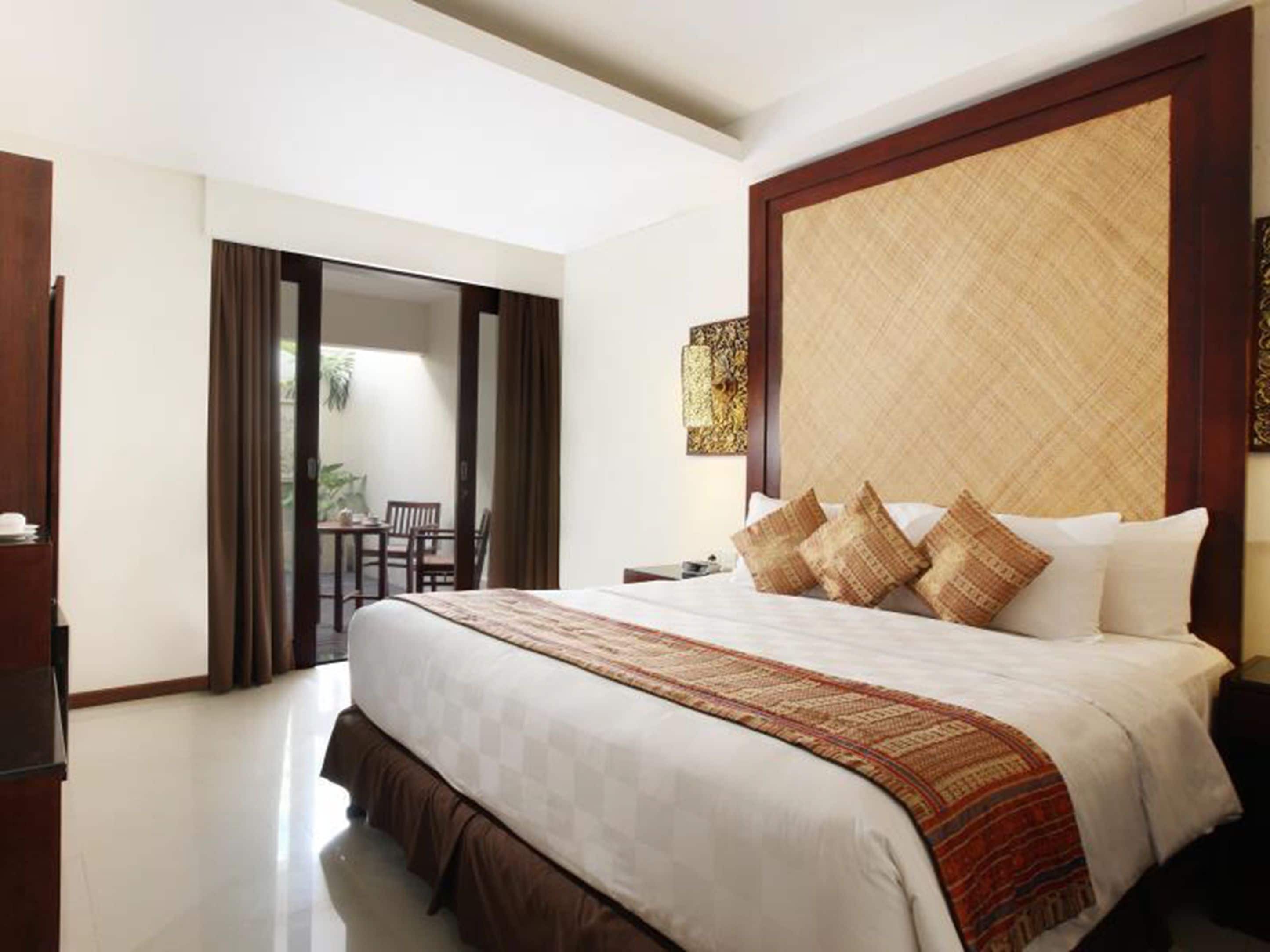 Une chambre d’hôtel avec un grand lit, une tête de lit en bois, une petite table et une vue sur un espace extérieur aménagé avec une table et des chaises.