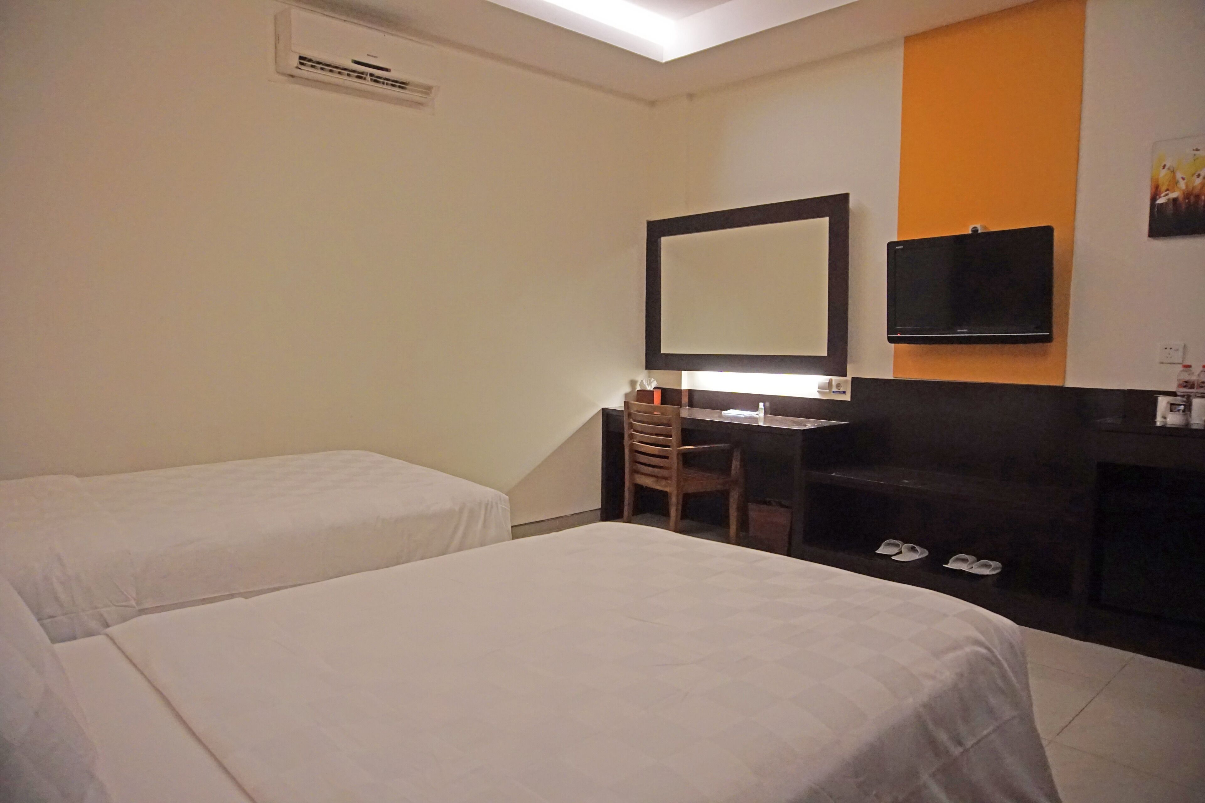 A hotel room with two beds, a desk with a chair, a flat-screen TV, and a small seating area with a bench and slippers.