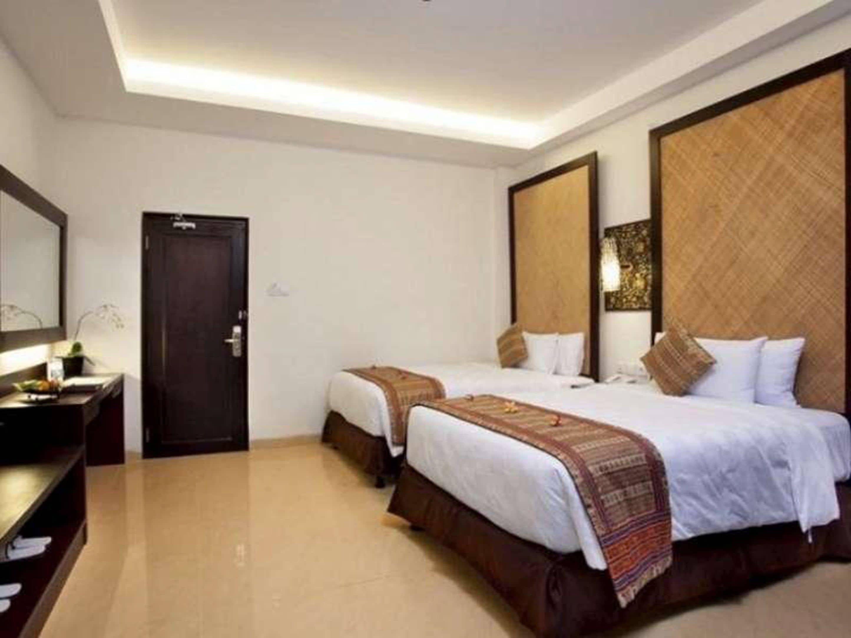 A hotel room with two beds, a dark wooden door, a small table with a lamp, and a large mirror on the wall.
