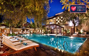A swimming pool with lounge chairs, a covered seating area, and a building with a checkmark indicating CHSE certification. - Best Western Kuta Villa (Tuban)