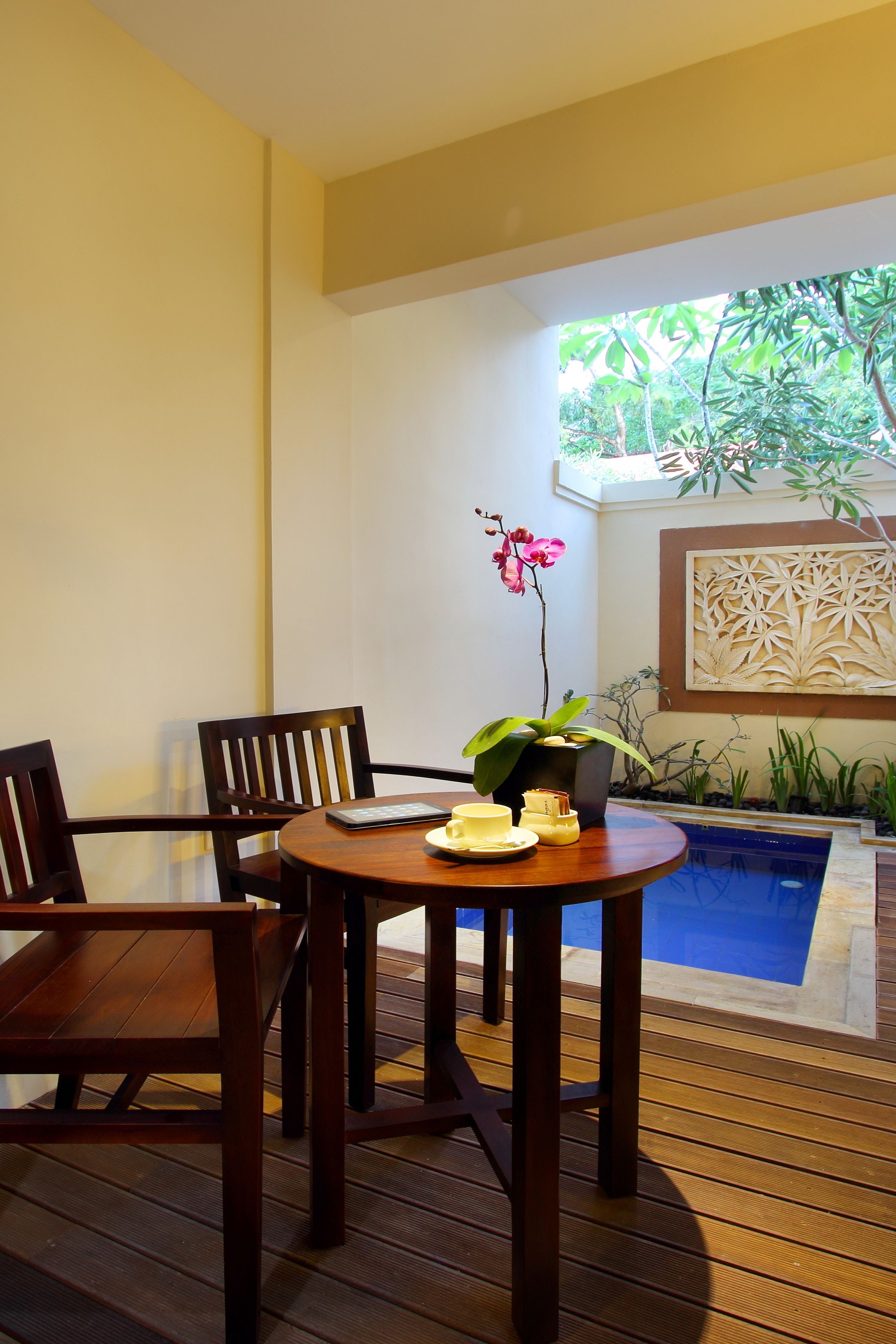 A wooden deck with a round table and chairs, a small indoor pool, and a potted plant with pink flowers.