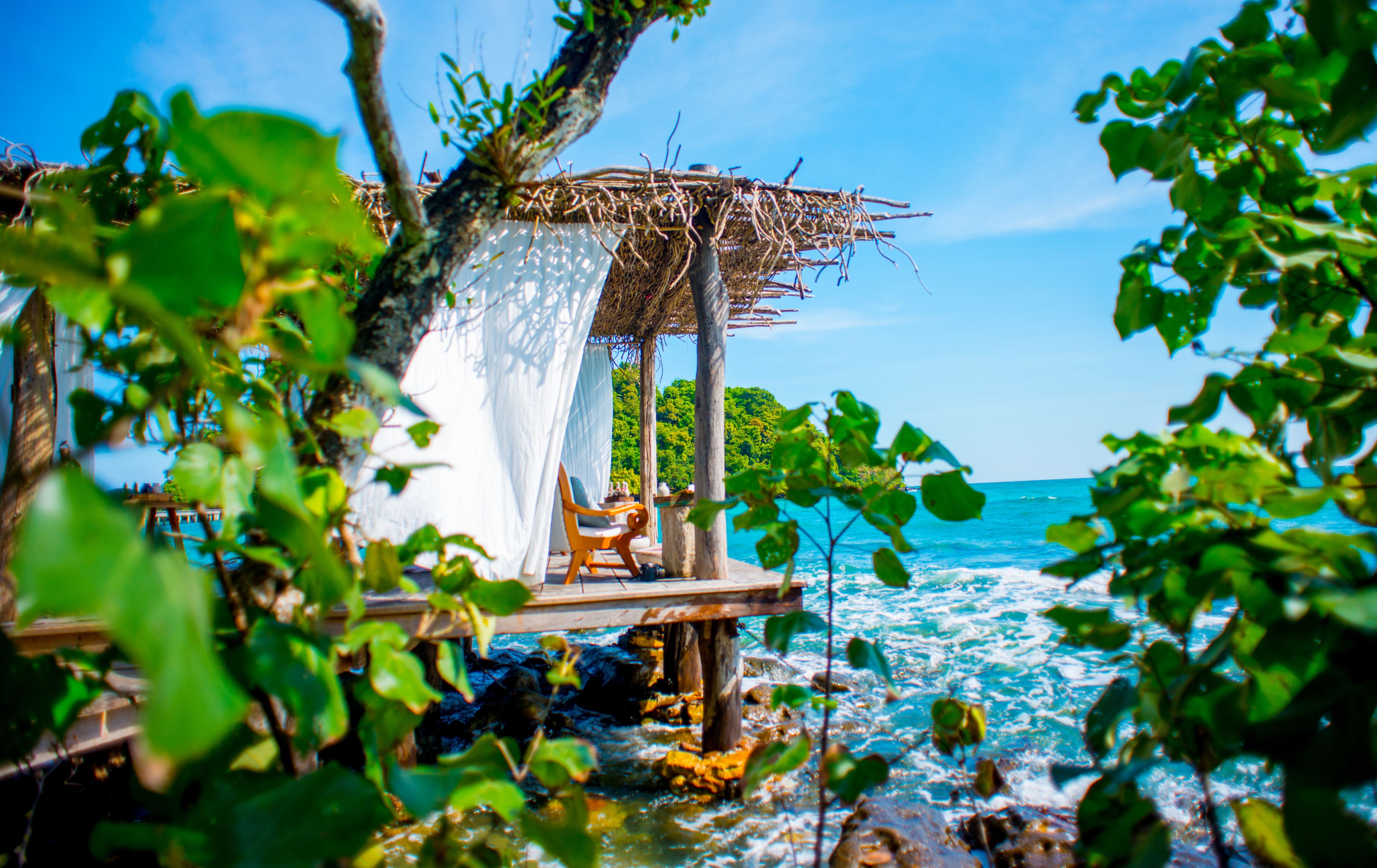 A wooden deck with a thatched roof and white curtains, overlooking a turquoise sea.