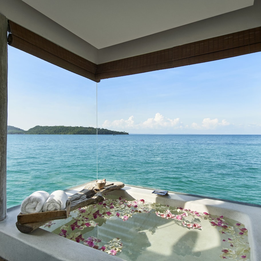 A room with a large window overlooking turquoise waters and a distant island. Inside, there's a wooden bench with towels and a wooden bowl.