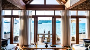 A room with large windows overlooking a body of water, wooden beams, and a view of a dock with two lanterns. - Song Saa Private Island (Koh Ouen)