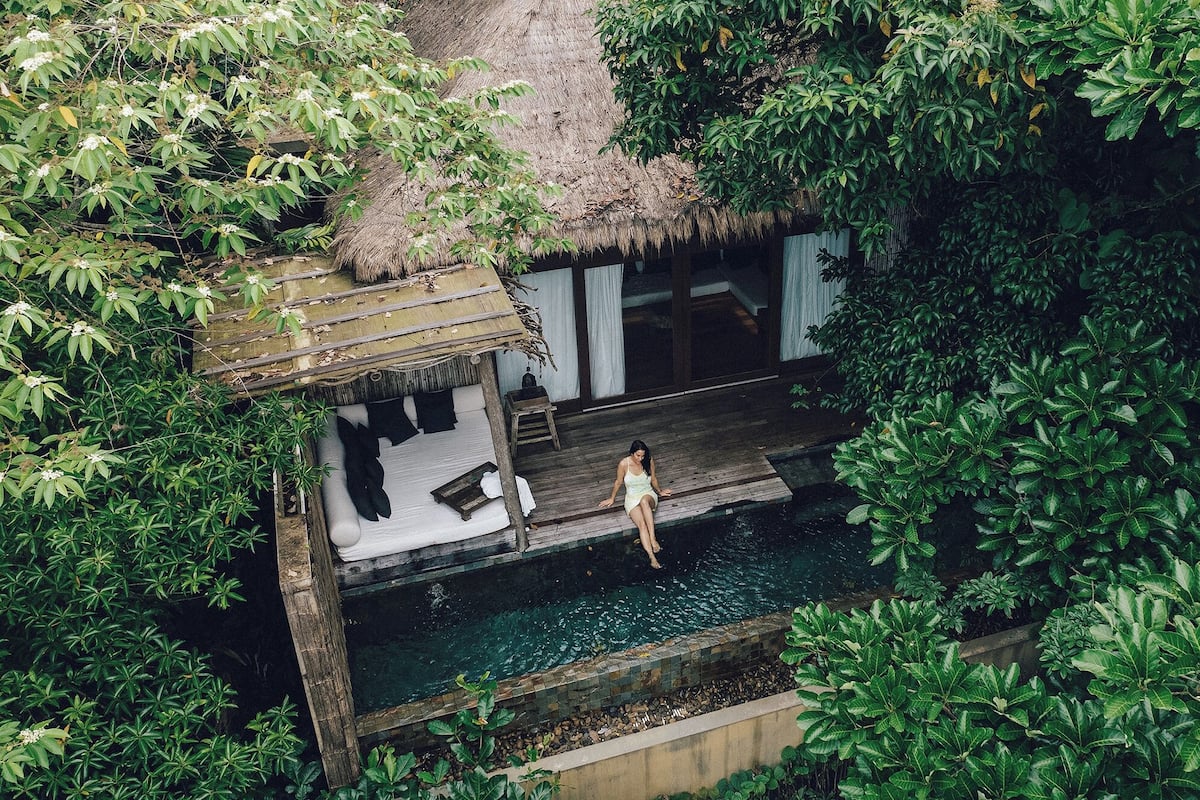 A person sitting on a wooden deck overlooking a pool, surrounded by lush greenery and a thatched-roof structure.