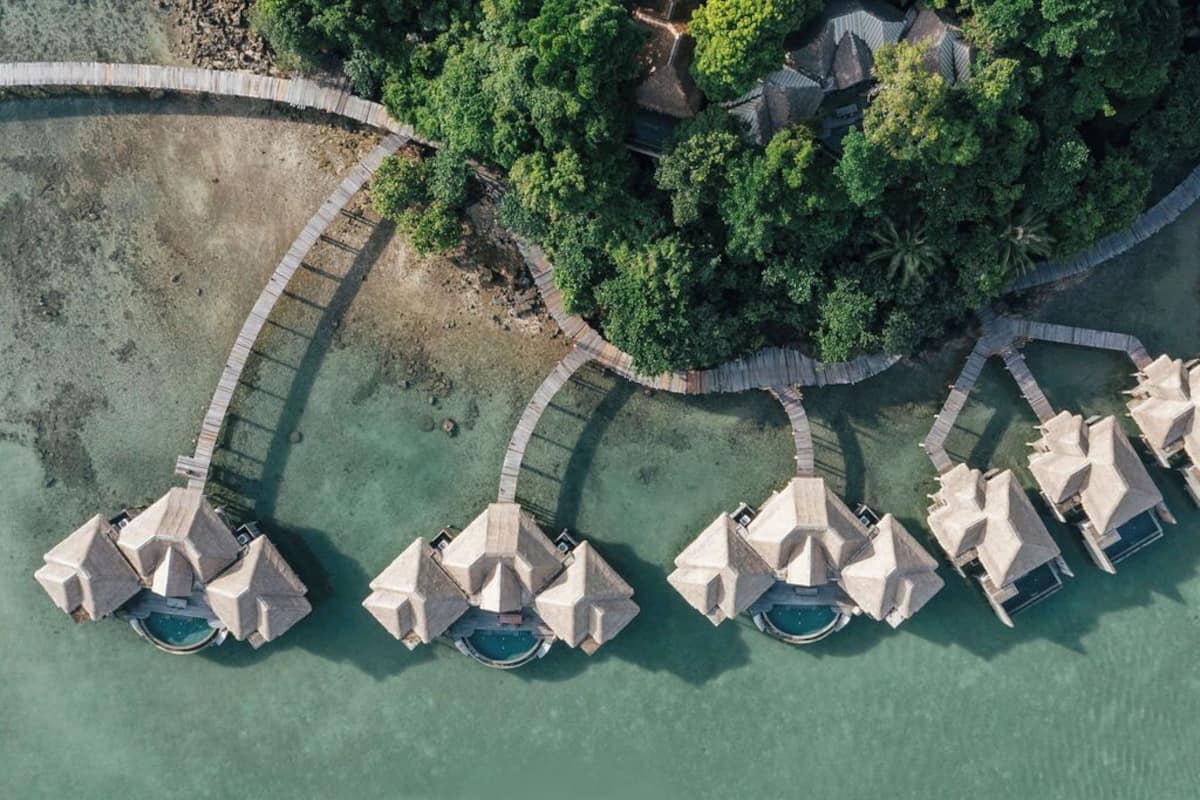 An aerial view of a resort with multiple overwater bungalows connected by a curved walkway, surrounded by lush greenery and clear turquoise waters.