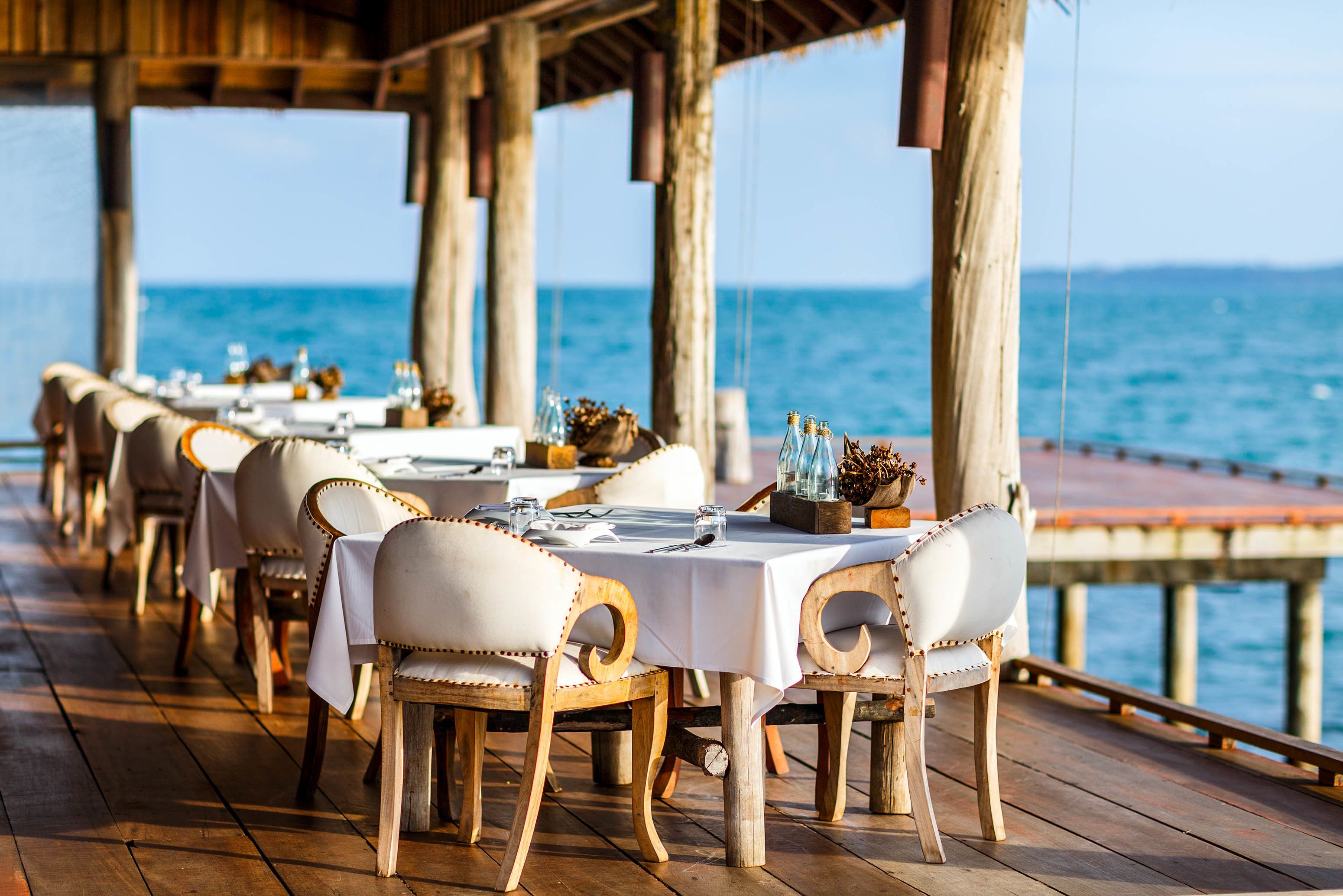 Un restaurante frente al mar, con mesas y sillas de madera, y el océano de fondo.