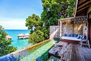 Um deck de madeira com sofá e mesa de jantar, com vista para uma piscina azul límpida e o oceano.