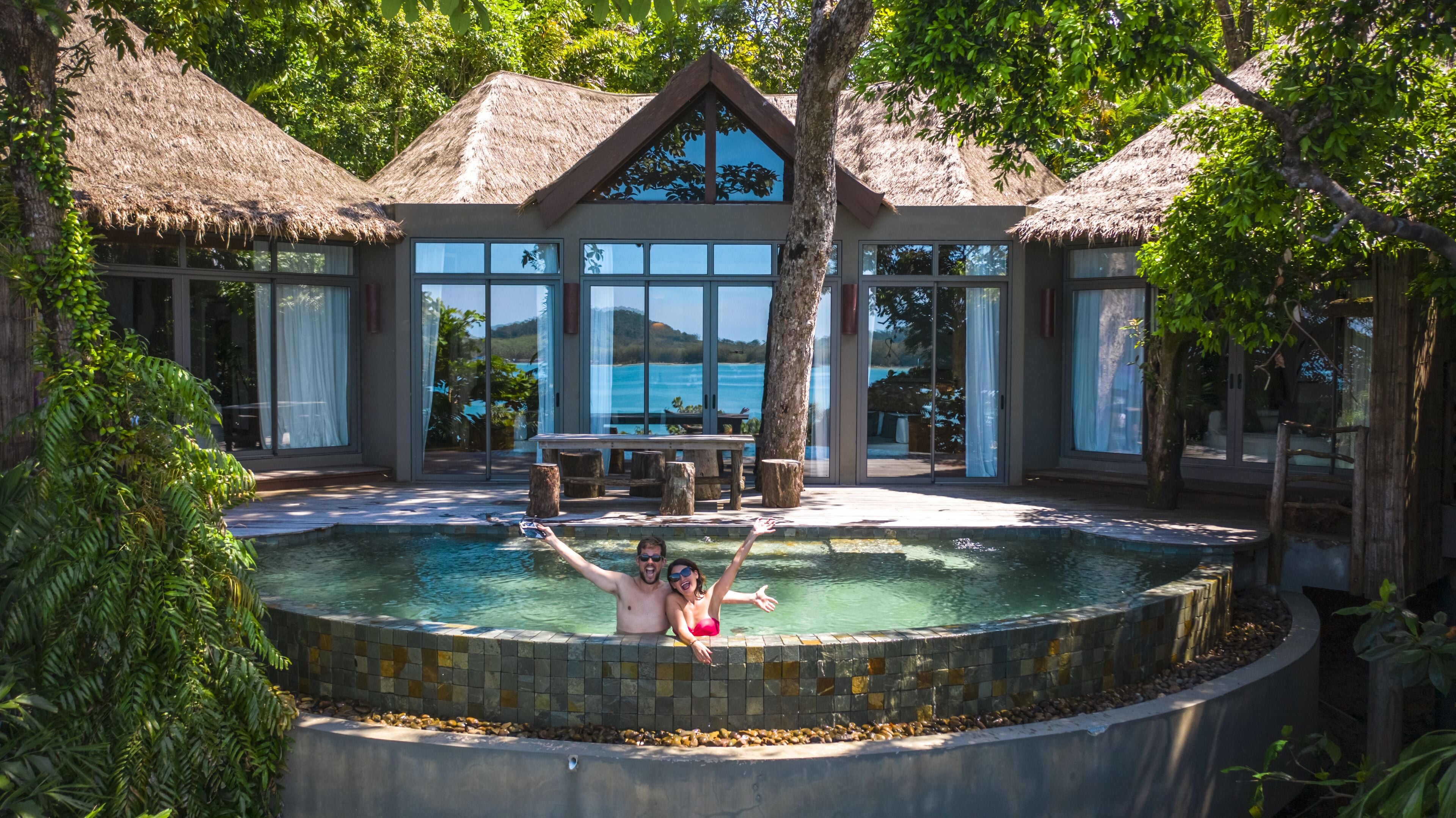 A swimming pool with a tiled edge, surrounded by lush greenery and a thatched-roof building.