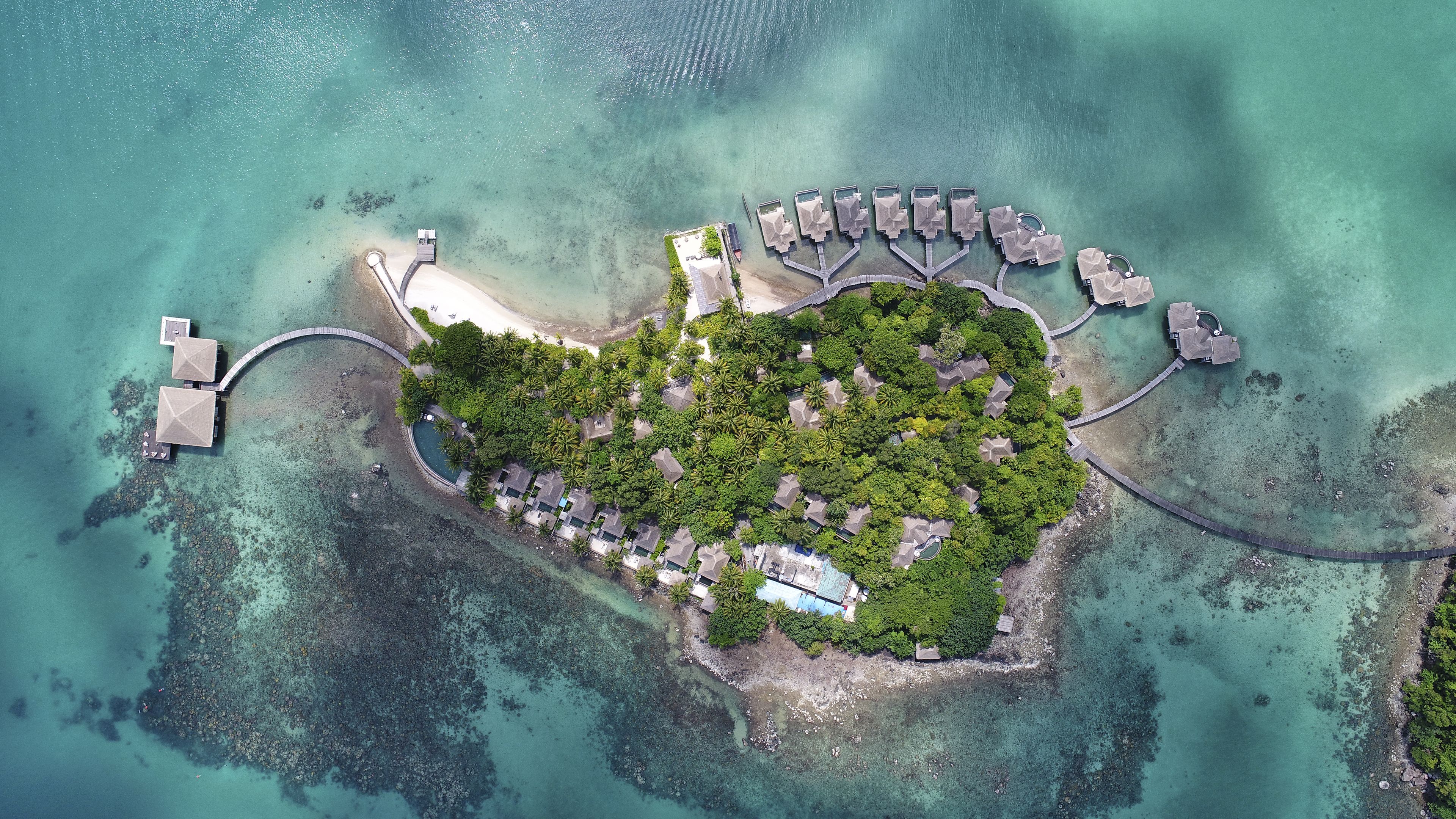 Vista aérea de un resort en una isla tropical con bungalows sobre el agua, un edificio central y una playa.