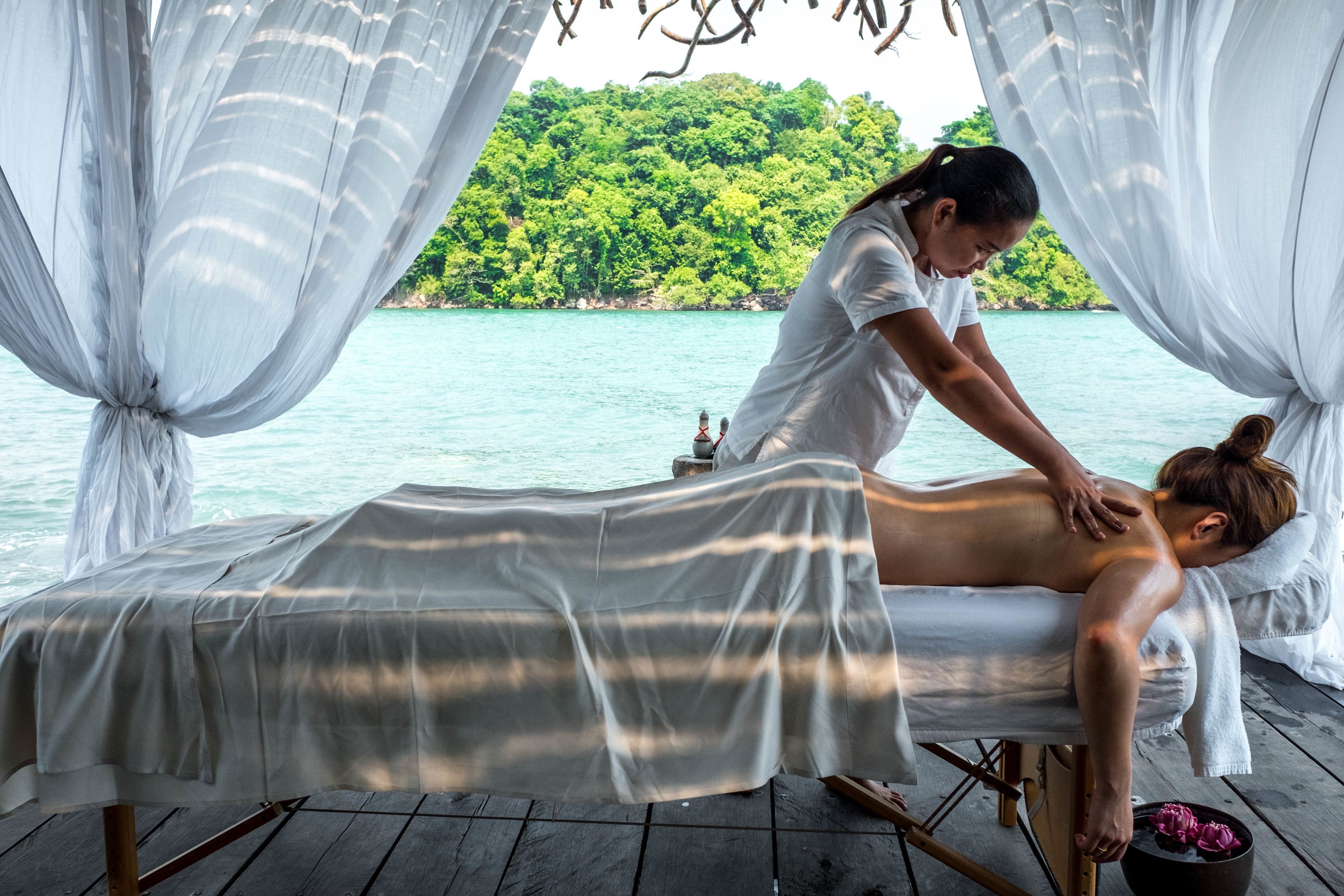 A massage therapy session on a wooden deck with a view of a turquoise sea and green hills.