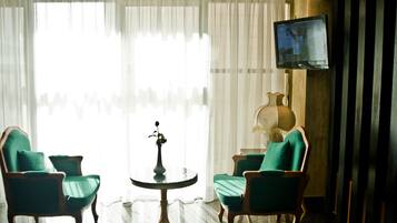 Ein Zimmer mit zwei grünen Polstersesseln, einem kleinen runden Tisch mit einer Vase und einem an der Wand montierten Fernseher.