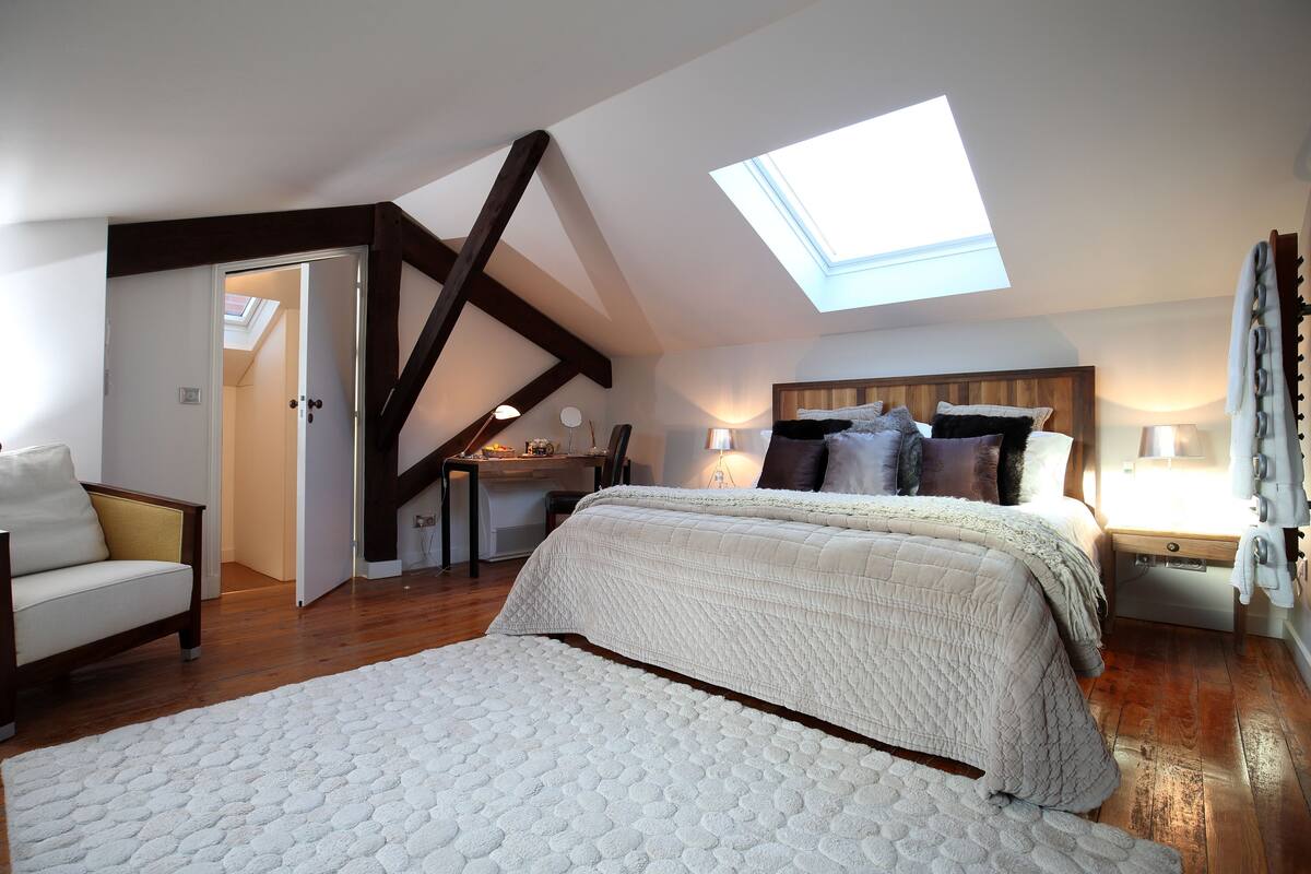 A bedroom with a skylight, a bed with a white quilt, a chair, and a desk with a lamp.