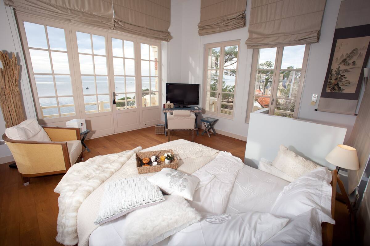 A spacious bedroom with a large bed, a desk with a TV, and a view of the sea.