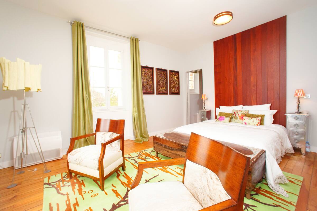 A bedroom with a bed, two chairs, a floor lamp, a dresser, and a window with curtains.