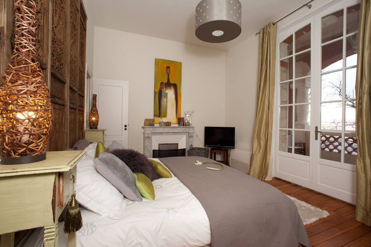 A bedroom with a bed, a television, a fireplace, and a large window with curtains.