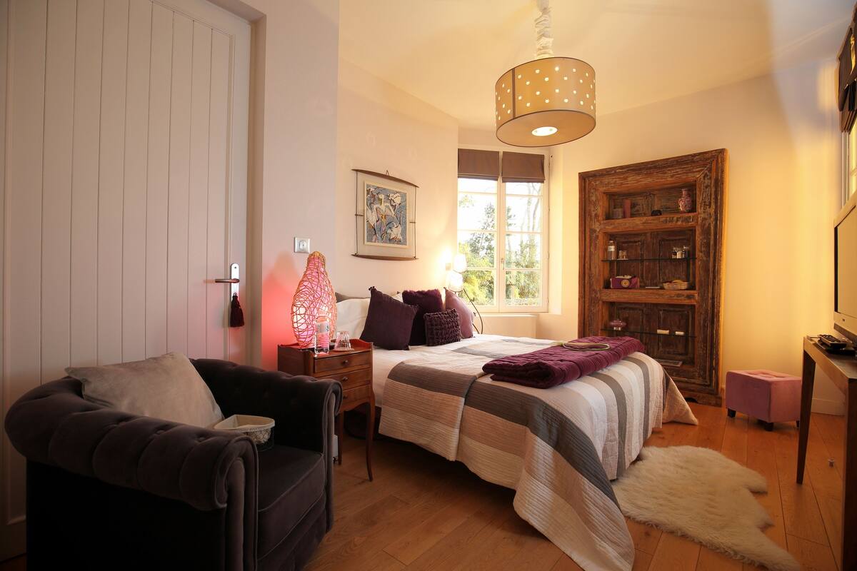 A bedroom with a bed, a nightstand, a chair, a window, a wooden cabinet, and a hanging light fixture.