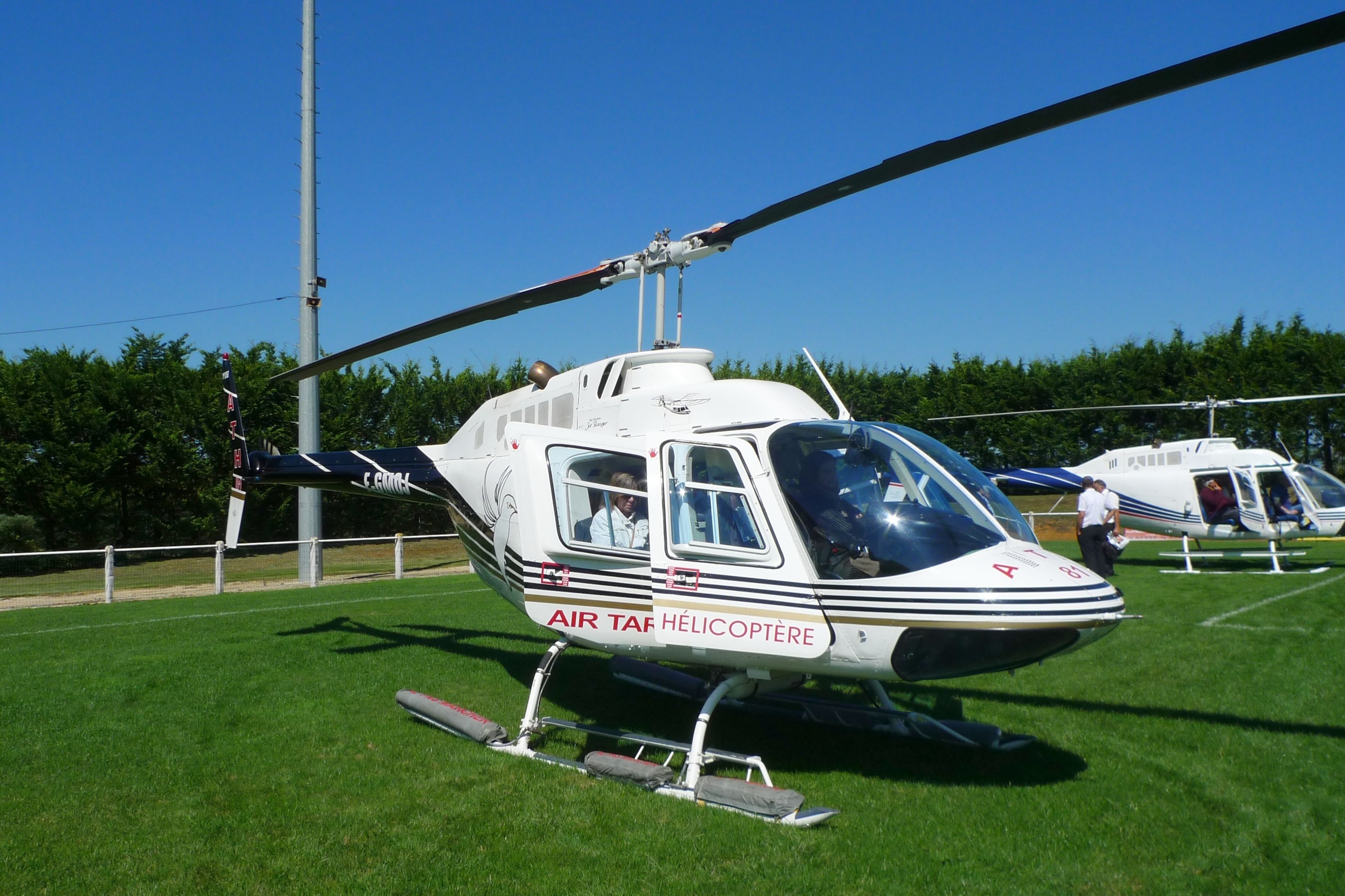 Een witte helikopter met daarop 'AIR TAF HÉLICOPTERE', geparkeerd op gras met een helderblauwe lucht op de achtergrond.