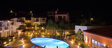 Vista nocturna de un resort con piscina, mobiliario exterior y varios edificios con balcones.