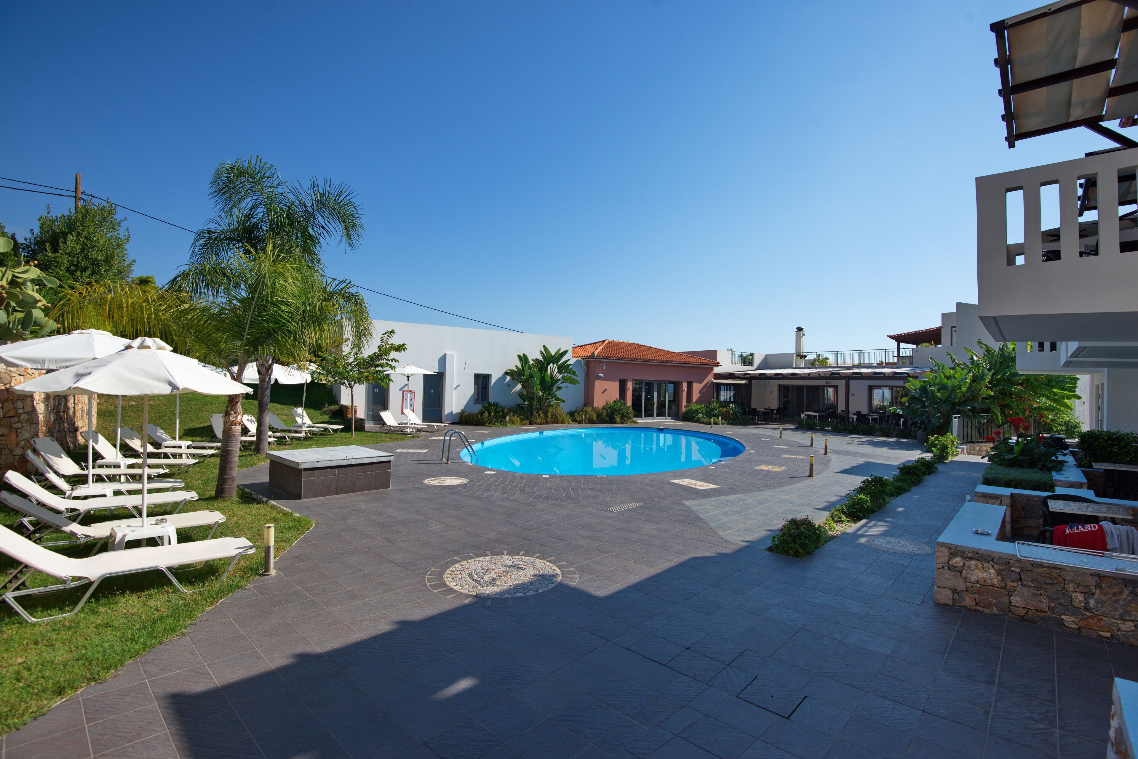 Un área de piscina con sillones de descanso y sombrillas, rodeada de un camino pavimentado y vegetación.