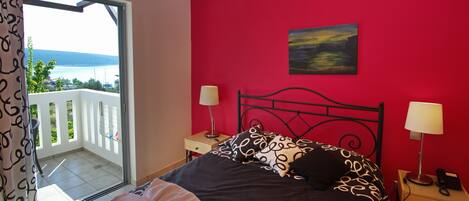A bedroom with a bed, two bedside tables, a painting, and a balcony with a view of the sea.