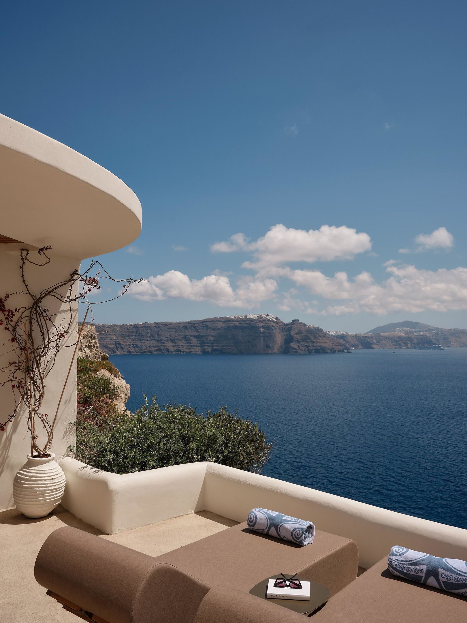 A balcony with a view of the sea and cliffs, featuring a lounge area with cushions and a book.