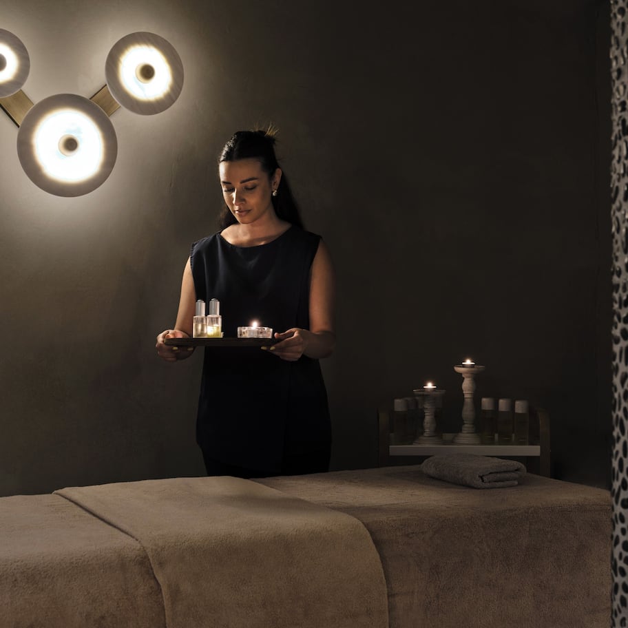 A person holding a tray with candles and bottles in a dimly lit room with a massage table and a decorative wall.