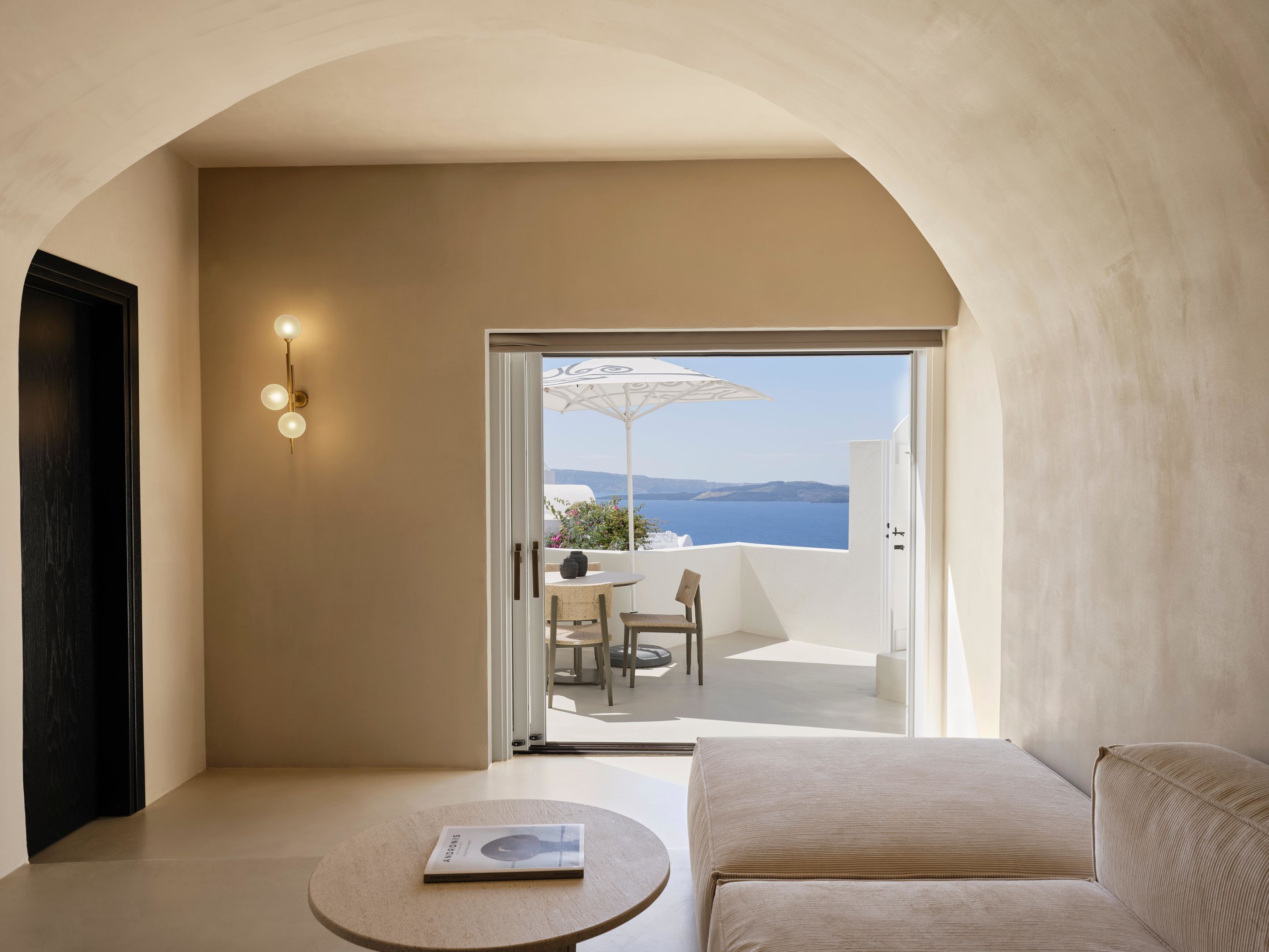 A modern room with a sofa, a small table with a book, and a view of a balcony with a table and chairs.