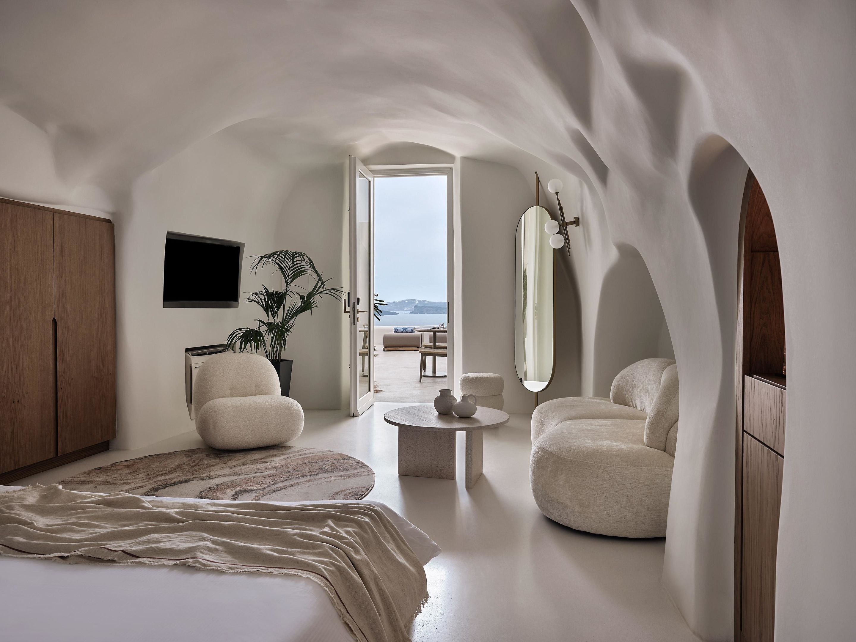 Un dormitorio moderno y minimalista con una cama grande, una zona de estar con una mesita de centro redonda y vistas al mar a través de un arco abierto.