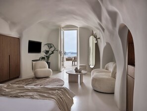 A modern, minimalist bedroom with a large bed, a seating area with a round coffee table, and a view of the sea through an open doorway. - Andronis Boutique Hotel (Santorini)