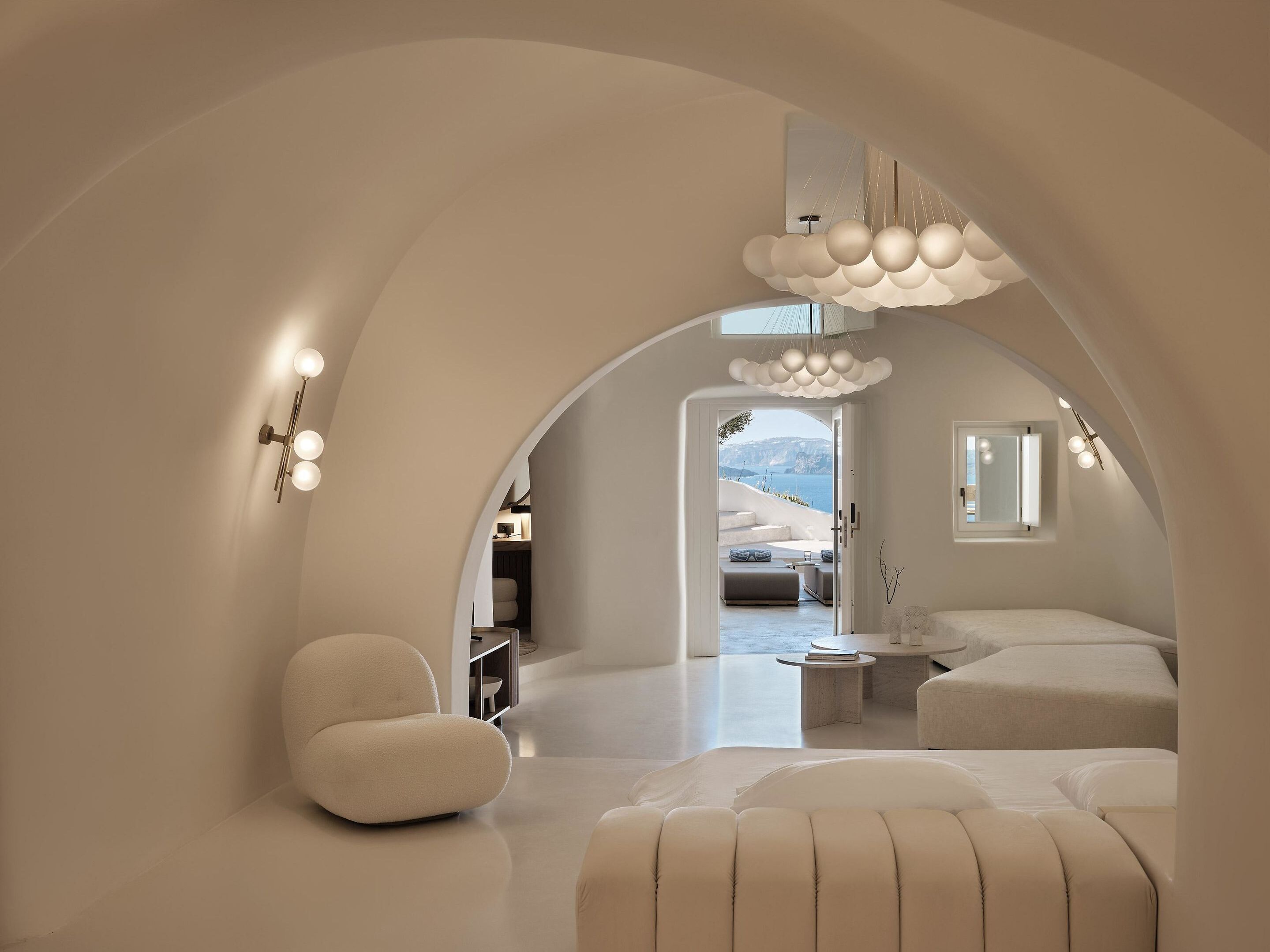 A modern interior with a curved ceiling, a seating area with a sofa and armchairs, a small table, and a view of a mountainous landscape through an open doorway.