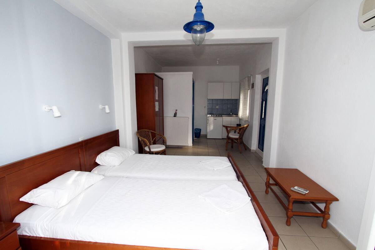 A hotel room with a double bed, a small wooden table, and a kitchenette in the background.